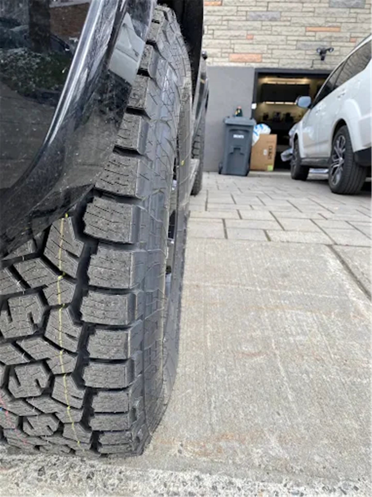 Close-up of all-terrain tire tread mounted on a vehicle near a driveway and garage.