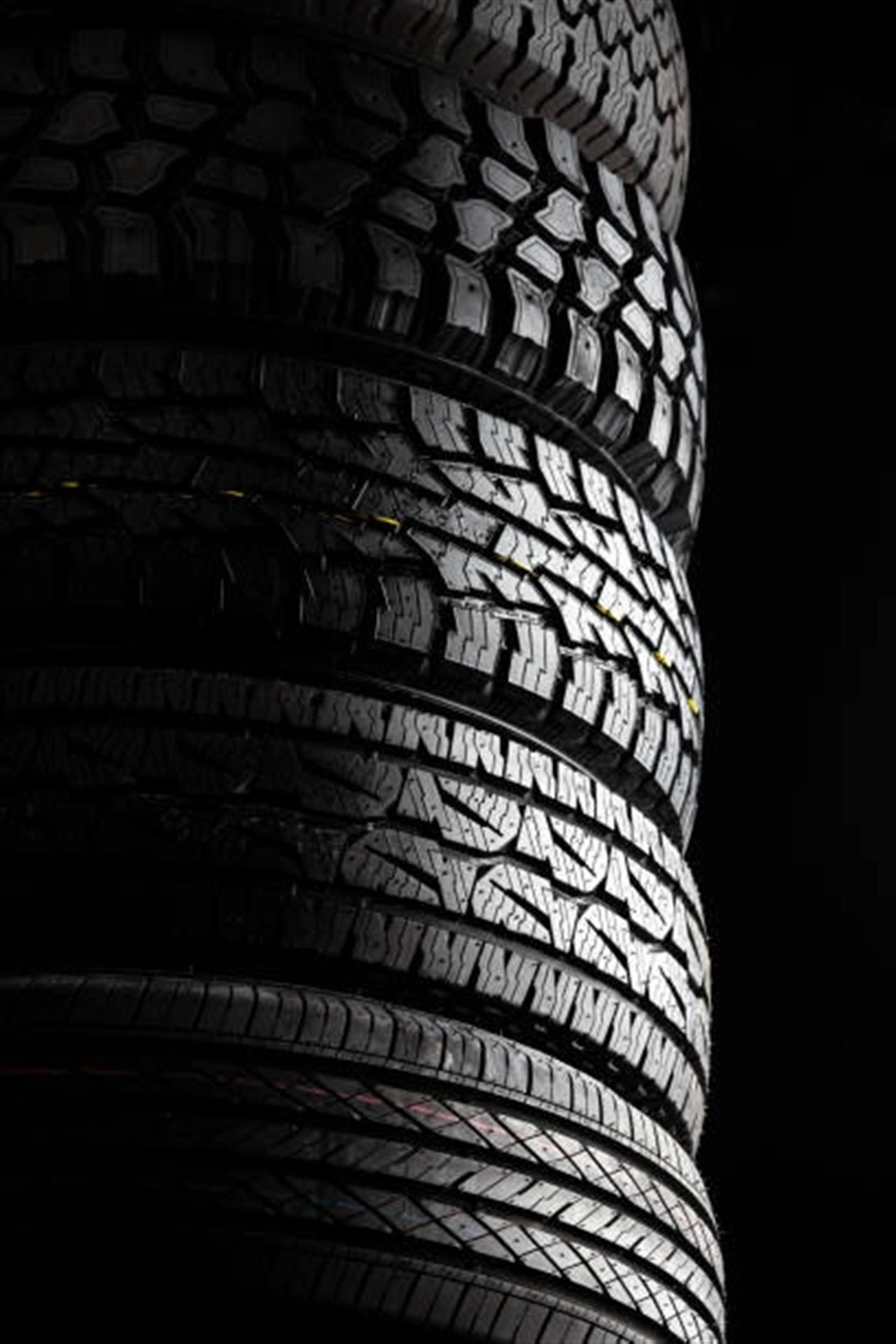 A close-up view of a stack of four car tires with different tread patterns, dramatically lit from the side against a black background.
